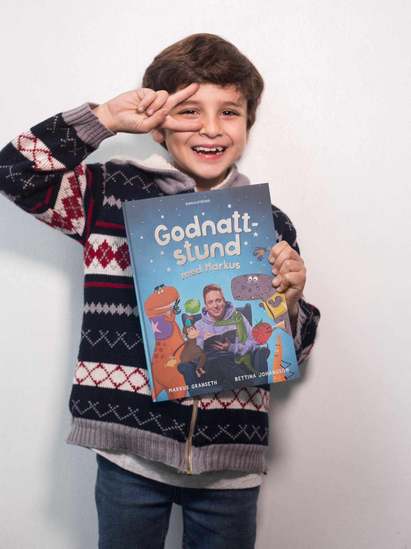 Ett skrattande barn håller boken Godnattstund med Markus, med illustrerade figurer på omslaget, och gör ett peace-tecken